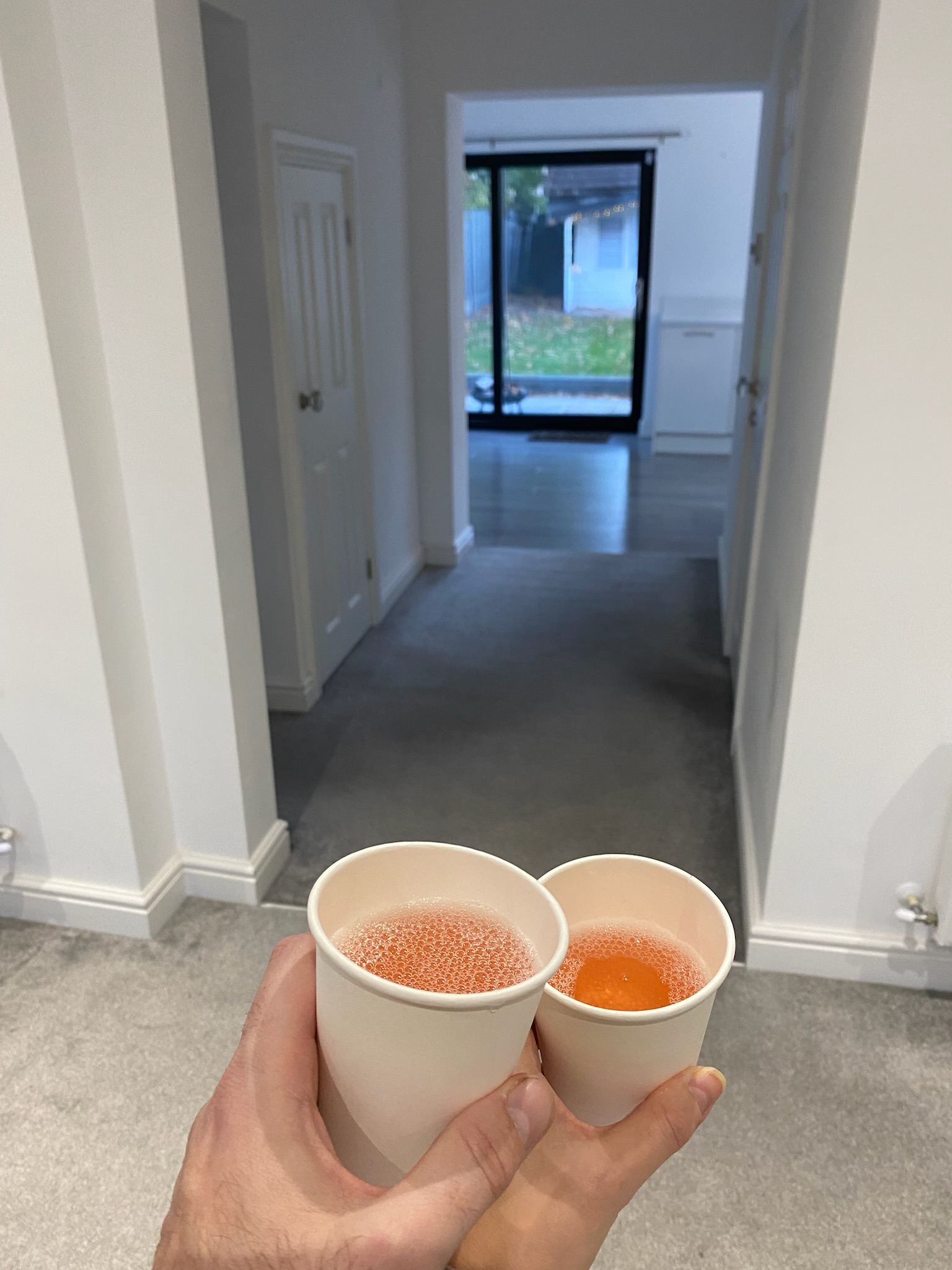 Two hands holding up paper cups filled with champagne in an empty house.
