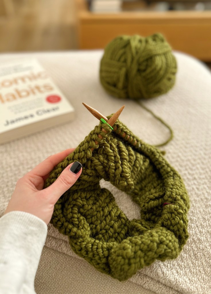 The neckline of a green sweater on knitting needles, placed on top of a cream sofa next to Atomic Habits by James Clear.